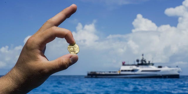 An explorer holds a gold coin found in the Bahamas where the Allen's exploration boat can be seen from afar.